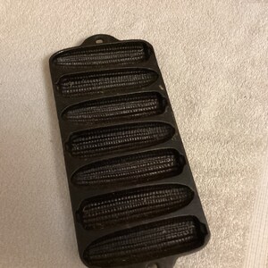 May include: Black cast iron cornbread pan with six individual molds. The pan has a handle on the top.