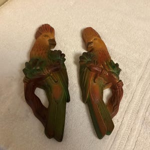 May include: Two wooden wall decor pieces shaped like parrots perched on branches. The parrots are painted in shades of brown, orange, green, and yellow.