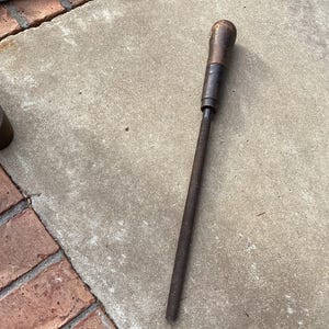 May include: A vintage metal tool with a wooden handle and a long, threaded shaft. The handle is dark brown, and the shaft is a dark, rusty color. The tool is lying on a concrete surface next to a brick border.
