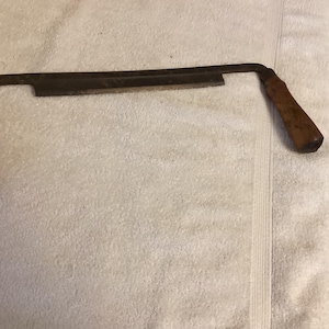 May include: A drawknife, a woodworking hand tool, is displayed. It features a long, straight blade with a cutting edge and two wooden handles. The blade is dark, and the handles are a warm brown.