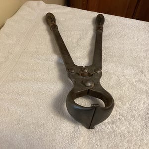 May include: An antique, dark metal farrier's tool, likely used for horseshoeing. The tool has two wooden handles and a large, curved jaw. The tool is resting on a white towel.
