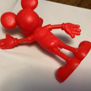 May include: A bright red, plastic Mickey Mouse figurine. The figure is lying on its side, with arms and legs outstretched. The iconic mouse has large, round ears, a round head, and large, red shoes. The figure is a classic toy.