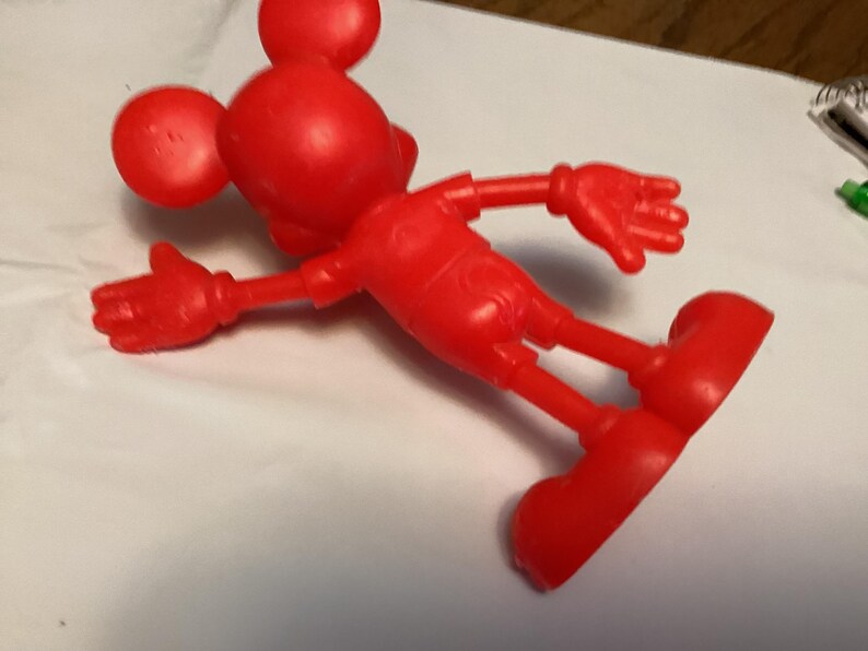 May include: A bright red, plastic Mickey Mouse figurine. The figure is lying on its side, with arms and legs outstretched. The iconic mouse has large, round ears, a round head, and large, red shoes. The figure is a classic toy.