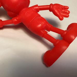 May include: A bright red, plastic figurine of a cartoon character, lying on its side. The figure has large, round ears, a short-sleeved shirt, shorts, and large, rounded shoes. The arms and legs are outstretched.