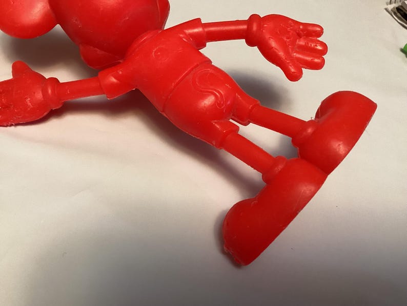May include: A bright red, plastic figurine of a cartoon character, lying on its side. The figure has large, round ears, a short-sleeved shirt, shorts, and large, rounded shoes. The arms and legs are outstretched.