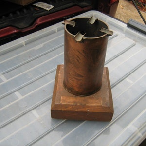May include: A vintage ashtray with a copper-colored cylindrical body and four cigarette rests. It is mounted on a wooden base with a copper plate. The ashtray is designed for cigars or cigarettes.