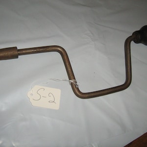 May include: A vintage, bronze-colored lug wrench with a rounded handle and a square end. The wrench has a curved design and a tag attached with the handwritten text "S-2".