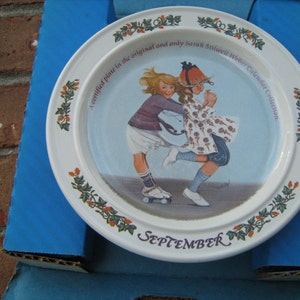 May include: A white ceramic plate with a green and orange floral border. The center of the plate features a painting of two girls on roller skates. The text "A certified plate in the original and only Sarah Stittwell Weber Calendar Collection" and "SEPTEMBER" are printed on the plate.