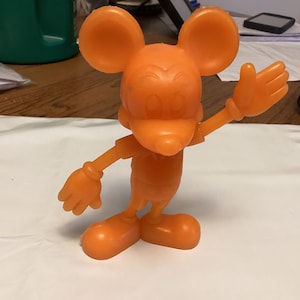 May include: An orange plastic figurine of a cartoon mouse with large ears, a smiling face, and gloved hands. The mouse is posed with one arm raised in a waving gesture. The figurine is standing on a white surface.