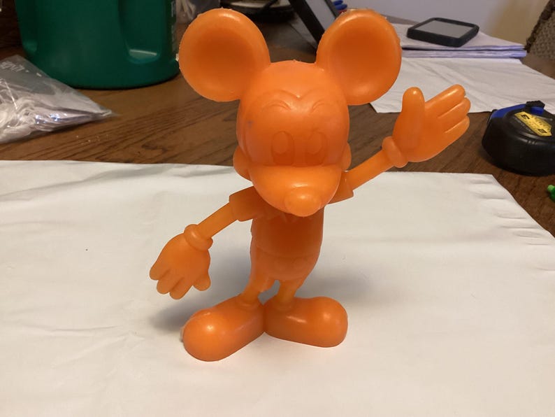 May include: An orange plastic figurine of a cartoon mouse with large ears, a smiling face, and gloved hands. The mouse is posed with one arm raised in a waving gesture. The figurine is standing on a white surface.