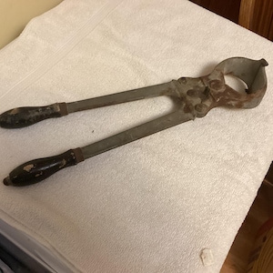 May include: An antique metal tool with long handles and a circular head. The handles have black wooden grips. The tool appears to be made of steel and shows signs of age and wear, with some rust visible.