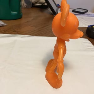May include: An orange, articulated toy figure of a cartoon character with large ears and a long snout. The figure is standing on a white surface, with a measuring tape and pen in the background. The toy is made of plastic.