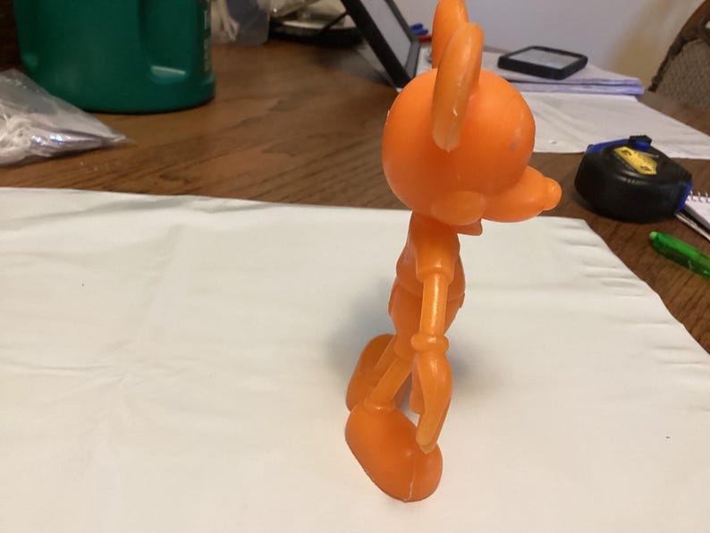 May include: An orange, articulated toy figure of a cartoon character with large ears and a long snout. The figure is standing on a white surface, with a measuring tape and pen in the background. The toy is made of plastic.