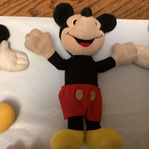 Puede incluir: Tres peluches de Mickey Mouse. Cada peluche tiene un cuerpo negro, orejas negras, una cara beige, guantes blancos, pantalones cortos rojos y zapatos amarillos. Mickey en el centro tiene los brazos extendidos en una pose de bienvenida.