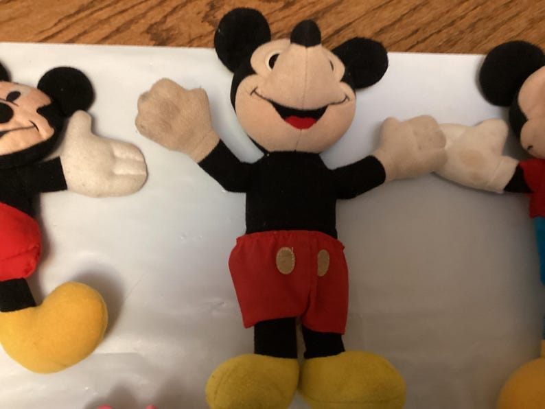 Puede incluir: Tres peluches de Mickey Mouse. Cada peluche tiene un cuerpo negro, orejas negras, una cara beige, guantes blancos, pantalones cortos rojos y zapatos amarillos. Mickey en el centro tiene los brazos extendidos en una pose de bienvenida.