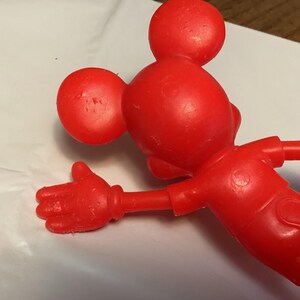 May include: A red plastic Mickey Mouse figurine, viewed from the back and side, with outstretched arm and hand. The figure has large round ears and a round head. The figure is on a white surface.