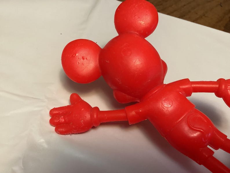 May include: A red plastic Mickey Mouse figurine, viewed from the back and side, with outstretched arm and hand. The figure has large round ears and a round head. The figure is on a white surface.