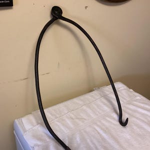 May include: A black metal hook with a rounded top and a hook at the bottom. The hook is hanging on a white wall.