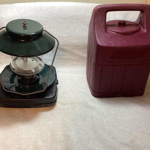 May include: A dark green lantern with a clear glass globe and a black base, positioned next to a maroon plastic Coleman carrying case. The lantern features a metal wire frame and a circular top hood, while the case has a molded handle and logo.