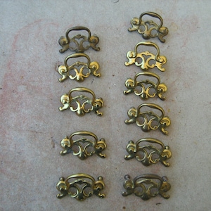 May include: A set of 12 antique brass drawer pulls with ornate designs. The pulls are all the same size and shape, and they have a slightly tarnished finish.