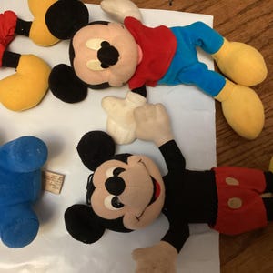 Puede incluir: Se muestran varios peluches vintage de Mickey Mouse. Los juguetes presentan las ic&oacute;nicas orejas negras de Mickey, pantalones cortos rojos, zapatos amarillos y guantes blancos. Un juguete es azul. Los juguetes est&aacute;n sobre una superficie blanca.