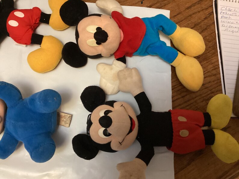 Puede incluir: Se muestran varios peluches vintage de Mickey Mouse. Los juguetes presentan las ic&oacute;nicas orejas negras de Mickey, pantalones cortos rojos, zapatos amarillos y guantes blancos. Un juguete es azul. Los juguetes est&aacute;n sobre una superficie blanca.