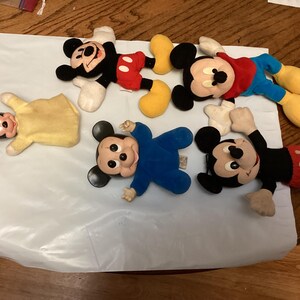 Puede incluir: Una colecci&oacute;n de peluches vintage de Mickey Mouse y Minnie Mouse. Los juguetes presentan dise&ntilde;os cl&aacute;sicos con colores negro, rojo, amarillo y azul. Los peluches est&aacute;n sobre una superficie blanca.