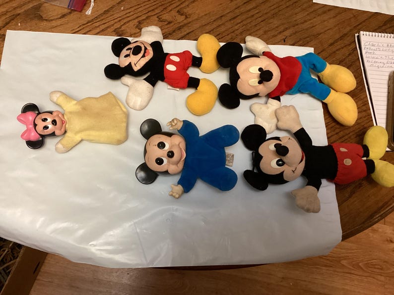 Puede incluir: Una colecci&oacute;n de peluches vintage de Mickey Mouse y Minnie Mouse. Los juguetes presentan dise&ntilde;os cl&aacute;sicos con colores negro, rojo, amarillo y azul. Los peluches est&aacute;n sobre una superficie blanca.