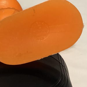 May include: Close-up of an orange plastic object with a rounded shape and a flat, oval base. The base has a circular marking. The object is partially obscuring a black, ridged surface.