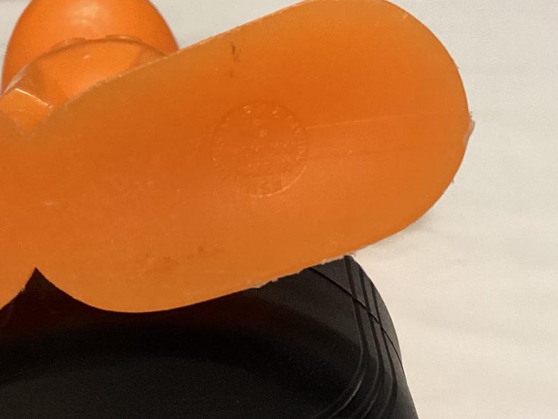 May include: Close-up of an orange plastic object with a rounded shape and a flat, oval base. The base has a circular marking. The object is partially obscuring a black, ridged surface.