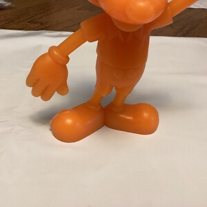 May include: Orange plastic figurine of a cartoon character with large feet and gloved hands. The figure is wearing a short-sleeved shirt and shorts. The figurine is standing on a white surface.