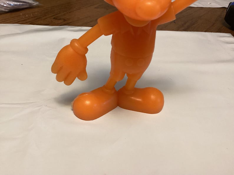 May include: Orange plastic figurine of a cartoon character with large feet and gloved hands. The figure is wearing a short-sleeved shirt and shorts. The figurine is standing on a white surface.