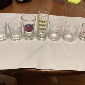May include: Seven clear glass shot glasses with various designs. The glasses are arranged in a row on a white surface. The designs include a red number 3, a gold design with the word "Tequila", and a blue and white design with the words "Universal Studios".