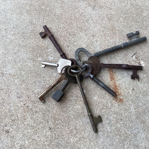 May include: A collection of antique keys with a variety of shapes and sizes, arranged in a starburst pattern on a gray concrete surface.