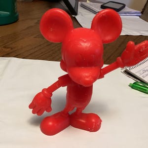 May include: A bright red Mickey Mouse figurine with outstretched arms and large ears. The vintage toy is posed on a white surface, with a notepad and pen in the background. The figurine is a classic Disney character.
