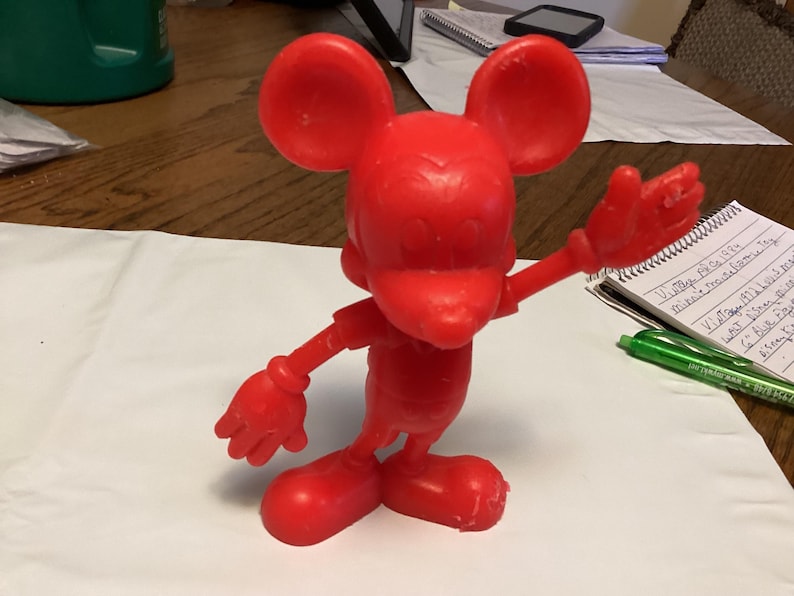 May include: A bright red Mickey Mouse figurine with outstretched arms and large ears. The vintage toy is posed on a white surface, with a notepad and pen in the background. The figurine is a classic Disney character.
