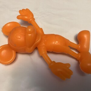May include: Orange plastic toy figure of a cartoon mouse, lying on its back. The figure has large round ears, a smiling face, and is wearing shorts and gloves. The arms and legs are outstretched.