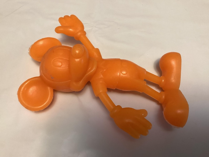 May include: Orange plastic toy figure of a cartoon mouse, lying on its back. The figure has large round ears, a smiling face, and is wearing shorts and gloves. The arms and legs are outstretched.
