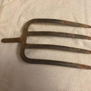 May include: An antique, rusty metal pitchfork with four tines. The handle and tines are a dark brown color with visible signs of age and wear. The pitchfork is designed for agricultural use.