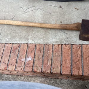 May include: A vintage axe with a long, light-colored wooden handle and a dark, rusty metal head. The axe is resting on a concrete surface with a brick border. The axe is a tool used for chopping wood.