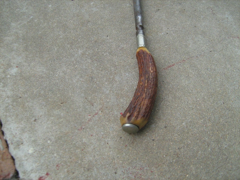 May include: A brown antler handle with a silver band and a metal cap on the end. The handle is attached to a metal rod.