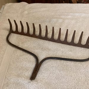 May include: A vintage garden rake with a curved handle and multiple tines. The metal rake is a rusty brown color and appears to be well-used. The tines are evenly spaced and curved. The rake is designed for gardening and yard work.