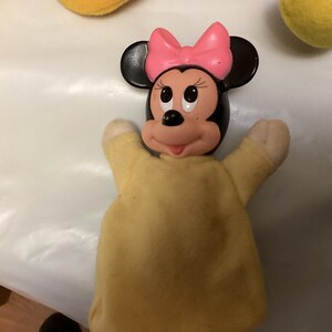Puede incluir: Una marioneta de mano de Minnie Mouse con un lazo rosa, orejas negras y un cuerpo amarillo. La cara de la marioneta es rosa y blanca con ojos negros y una nariz negra. Los brazos de la marioneta est&aacute;n levantados.