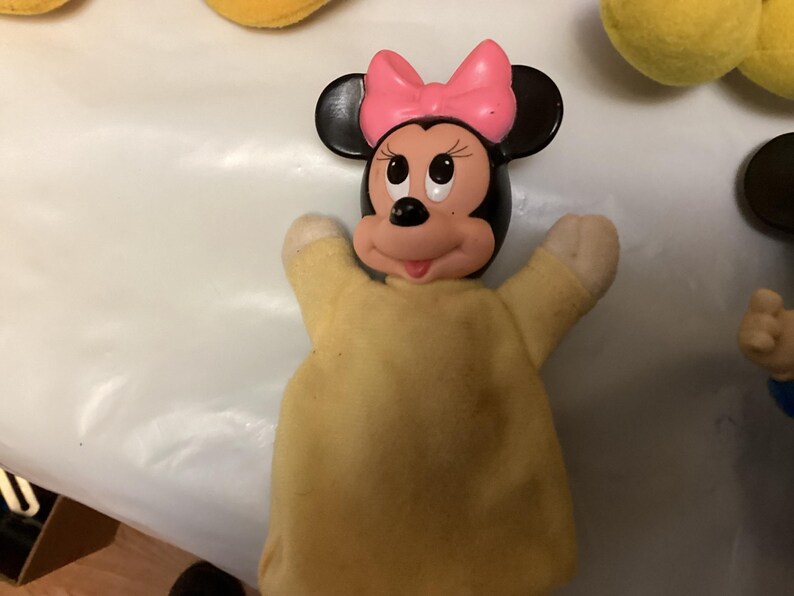 Puede incluir: Una marioneta de mano de Minnie Mouse con un lazo rosa, orejas negras y un cuerpo amarillo. La cara de la marioneta es rosa y blanca con ojos negros y una nariz negra. Los brazos de la marioneta est&aacute;n levantados.