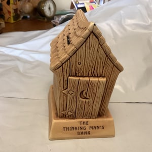 May include: Tan ceramic bank shaped like a small wooden outhouse. The roof has a shingle design, and the door features a crescent moon and star details. The base reads "THE THINKING MAN'S BANK".