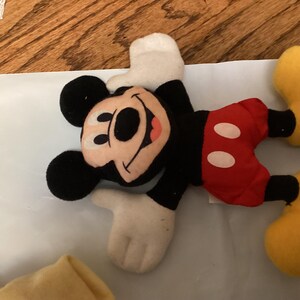 Puede incluir: Un peluche de Mickey Mouse tumbado. Mickey lleva pantalones cortos negros, pantalones rojos con botones blancos, zapatos amarillos y guantes blancos. Su cara es de color beige con nariz y orejas negras, y una boca roja sonriente.