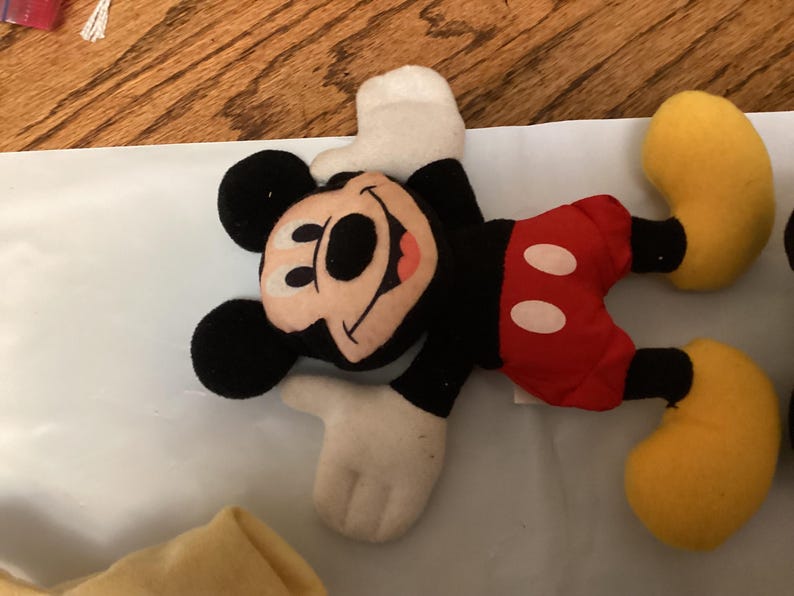 Puede incluir: Un peluche de Mickey Mouse tumbado. Mickey lleva pantalones cortos negros, pantalones rojos con botones blancos, zapatos amarillos y guantes blancos. Su cara es de color beige con nariz y orejas negras, y una boca roja sonriente.