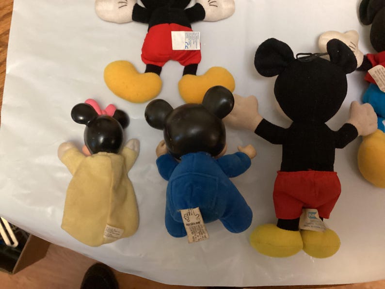 Puede incluir: Una colecci&oacute;n de mu&ntilde;ecos de peluche vintage de Mickey Mouse y Minnie Mouse. Los mu&ntilde;ecos de Mickey visten pantalones cortos rojos, cuerpos negros, zapatos amarillos y guantes blancos. Los mu&ntilde;ecos de Minnie visten vestidos amarillos y lazos rosas. Los mu&ntilde;ecos est&aacute;n sobre una superficie blanca.