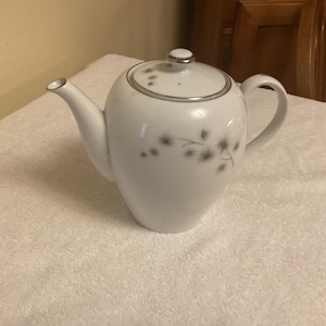 May include: A white porcelain teapot featuring a delicate gray dandelion seed head pattern on the body and lid. The teapot has a curved handle, a tapered spout, and a matching lid with a silver-toned rim accent along the top edge and spout.