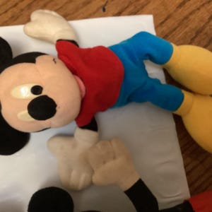 Puede incluir: Un peluche de Mickey Mouse, con una camisa roja, pantalones cortos azules, zapatos amarillos y guantes blancos. El ic&oacute;nico personaje tiene orejas negras y una cara bronceada con ojos grandes y redondos. El juguete est&aacute; sobre una superficie blanca.